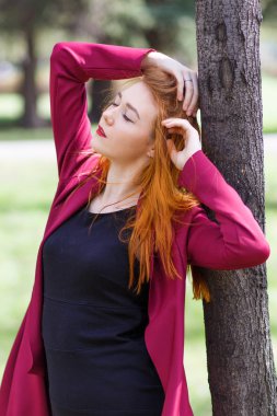 Kırmızı saçlı kız kırmızı giysiler içinde bir ağacın yanında poz.
