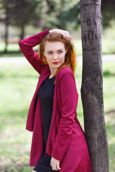 Chica pelirroja posando cerca de un árbol con ropa roja . — Foto de Stock