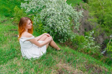 Beyaz elbiseli kızıl saçlı kız, çiçek açan bir Elma ağacının arka planında yeşil çimenlerin üzerinde oturuyor. Doğada kız bahar.
