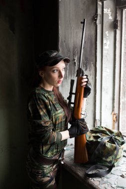 İnce kızıl saçlı bir kız kırık pencerede tüfekle duruyor. Yeşil kamuflajlı, silahlı kız. Kızlar için askerlik hizmeti. Kız orduda..
