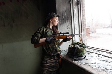 İnce kızıl saçlı bir kız kırık pencerede tüfekle duruyor. Yeşil kamuflajlı, silahlı kız. Kızlar için askerlik hizmeti. Kız orduda..