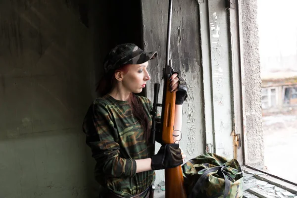 Тонкая рыжая девушка стоит у разбитого окна с винтовкой. Девушка в зеленом камуфляже с пистолетом. Военная служба для девочек. Девушка в армии. . — стоковое фото