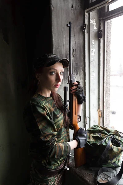 Тонкая рыжая девушка стоит у разбитого окна с винтовкой. Девушка в зеленом камуфляже с пистолетом. Военная служба для девочек. Девушка в армии. . — стоковое фото