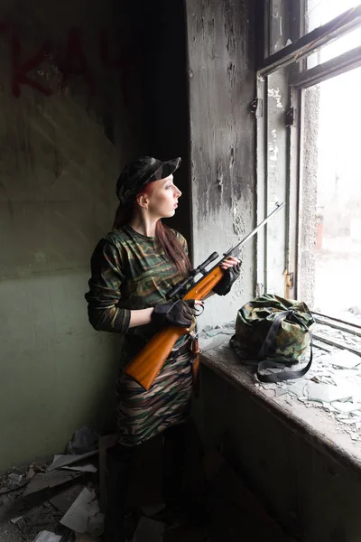 Uma menina ruiva fina fica na janela quebrada com uma espingarda. A rapariga de camuflagem verde com uma arma. Serviço militar para meninas. A rapariga está no exército. . — Fotografia de Stock