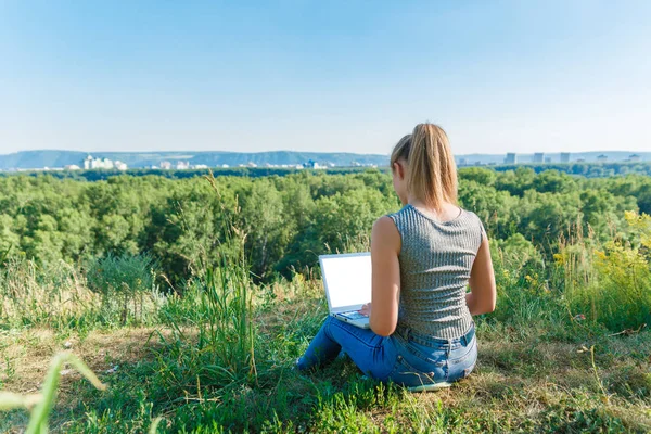 Girl with laptop on the nature on the background of forest and sky. The concept of combining leisure and work. Individual entrepreneur. Work at the computer away from the city.
