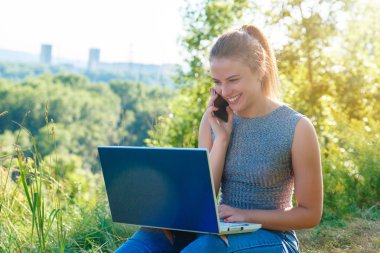 Kız bir akıllı telefon konuşuyor ve dizüstü monitörbakıyor. Bir dizüstü bilgisayar ile doğada kot pantolon kız. Tatildeyken iş yapmak.