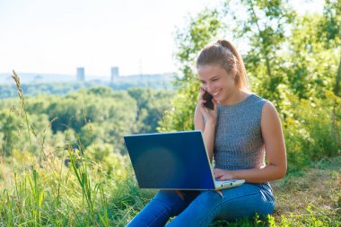 Kız bir akıllı telefon konuşuyor ve dizüstü monitörbakıyor. Bir dizüstü bilgisayar ile doğada kot pantolon kız. Tatildeyken iş yapmak.