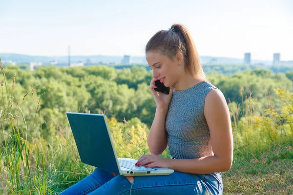 Το κορίτσι μιλάει σε ένα smartphone και κοιτάζοντας την οθόνη του φορητού υπολογιστή. Κορίτσι με τζιν στη φύση με ένα φορητό υπολογιστή. Επιχειρήσεις κατά τη διάρκεια των διακοπών. — Φωτογραφία Αρχείου