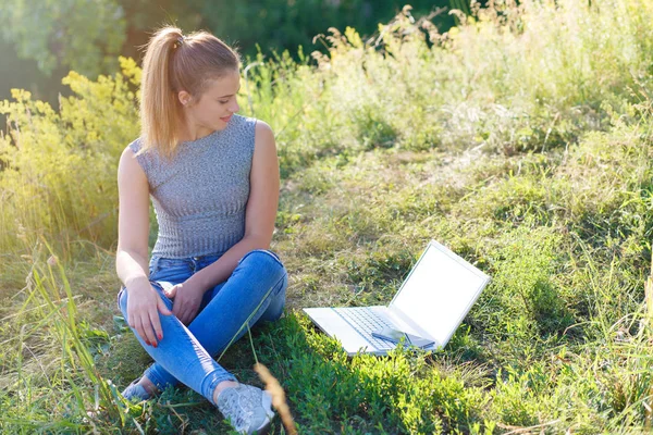 Νεαρό κορίτσι με τζιν στη φύση κάθεται στο έδαφος και κοιτάζοντας την οθόνη του φορητού υπολογιστή. Φορητός υπολογιστής με λευκή οθόνη. Χώρος αντιγραφής στον φορητό υπολογιστή σας. — Φωτογραφία Αρχείου