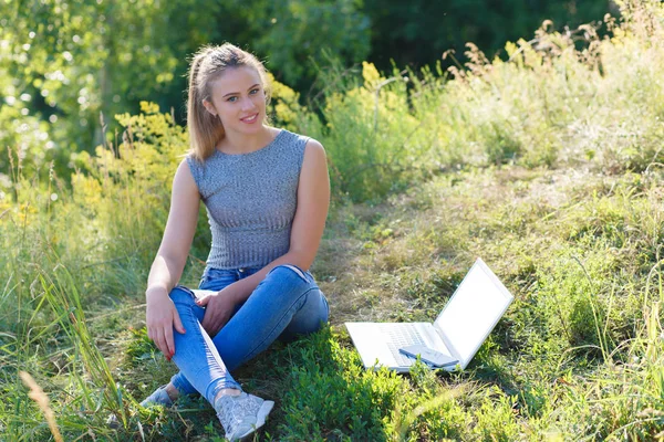 Νεαρό κορίτσι με τζιν στη φύση κάθεται στο έδαφος. Δίπλα στο κορίτσι είναι ένας φορητός υπολογιστής. Λευκή οθόνη στο φορητό υπολογιστή. — Φωτογραφία Αρχείου