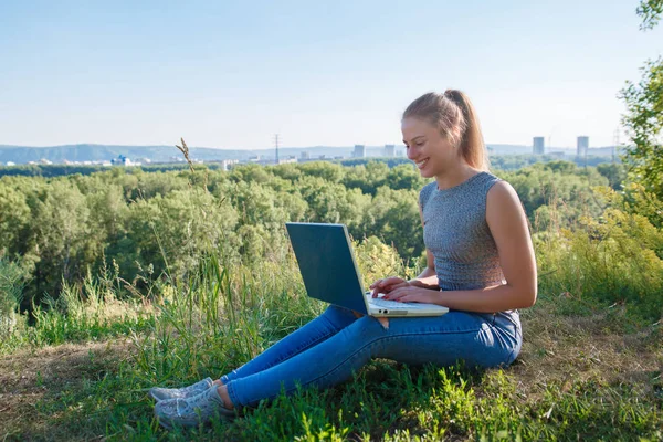 Νεαρό κορίτσι χαμογελά στη φύση με φορητό υπολογιστή στην αγκαλιά της. Το κορίτσι είναι χαρούμενος κοιτάζοντας την οθόνη του φορητού υπολογιστή στο φόντο του δάσους και της πόλης. — Φωτογραφία Αρχείου