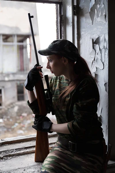 Uma jovem com uma espingarda de uniforme na janela a olhar para a rua. A mulher franco-atirador em um terno verde e boné . — Fotografia de Stock