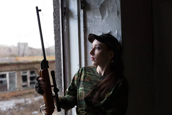 Молодая женщина в форме с винтовкой у окна смотрит на улицу. Женщина-снайпер в зеленом костюме и кепке . — стоковое фото