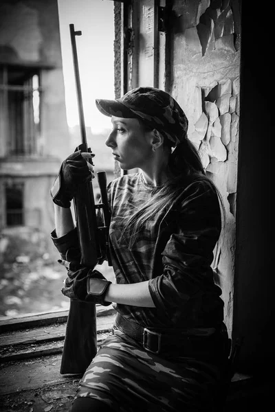 A young woman with a rifle in uniform at the window looking at the street. Female sniper in camouflage and cap. Black and white photo. — Stock Photo, Image