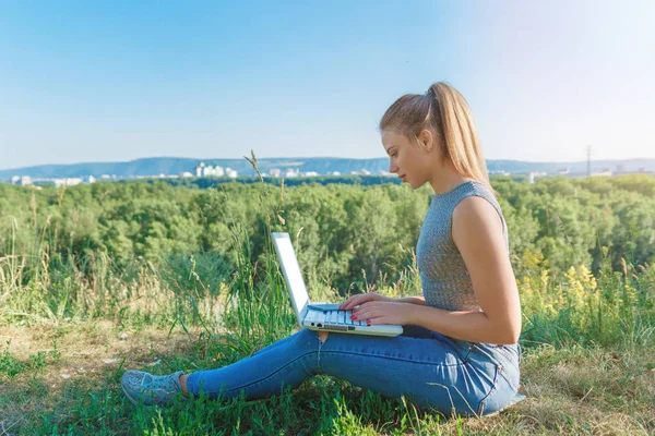 A girl with a laptop in nature, against the forest and the sky with sunlight. The concept of combining leisure and work. Individual entrepreneur. Work at the computer away from the city.