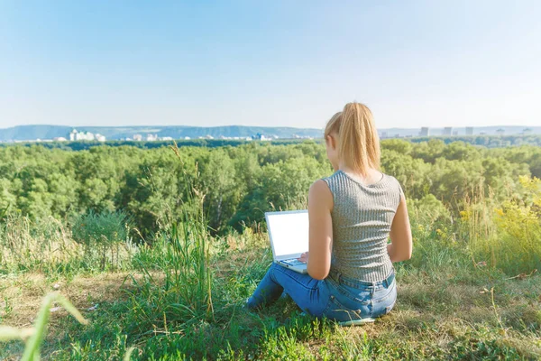 A girl with a laptop in nature, against the forest and the sky with sunlight. The concept of combining leisure and work. Individual entrepreneur. Work at the computer away from the city.