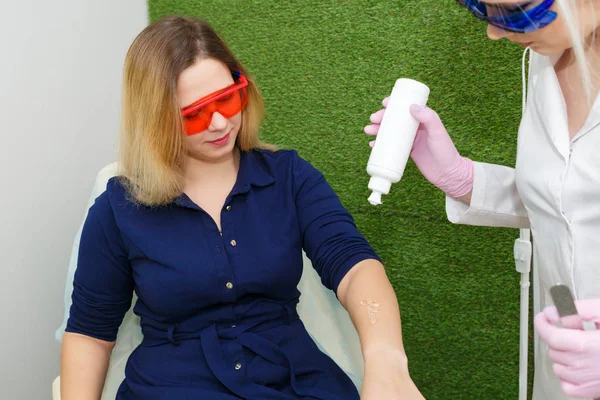 Una mujer joven se está preparando para la depilación láser. Una chica en un vestido azul y gafas está en el salón para eliminar el cabello con un láser . — Foto de Stock