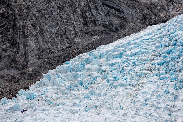 在左下角的徒步旅行者群增加了在蓝色冰上的尺度感 这是并列反对黑色灰色岩石在 Franz 约瑟夫冰川 南岛新西兰 — 图库照片