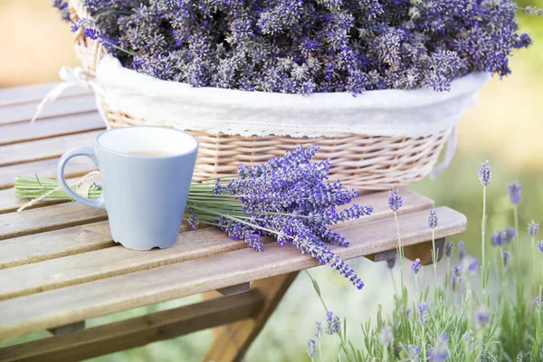 A basket filled with purple flowers stands on a wooden table on a background of green lavender fields. — Stock Photo, Image