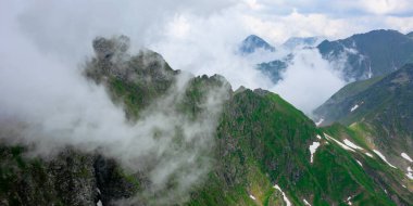 Bulutların arasındaki yüksek dağ zirveleri. Tepelerde karla kaplı harika bir yaz manzarası. Mavi gökyüzü bulutlu
