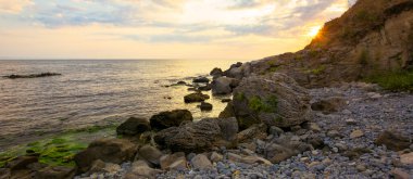 Taş ve çakıl taşlı deniz plajı. Güneşin doğuşunda gökyüzünde bulutlar olan güzel bir manzara. Bir körfezin geniş panoramik görüntüsü