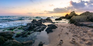 Okyanus kıyısında huzurlu bir gün batımı. Dalgalar kumlu sahilde kayalara çarpıyor. Ufkun üzerindeki güzel bulutlar