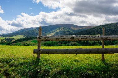 Kırsal bölgedeki tepede çit var. Karpat Dağları 'ndaki sonbahar manzarası. Gökyüzünde bulutlar olan güneşli bir hava. Tepeler uzaktaki dağ sırtında yuvarlanıyor.