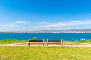 Güneşli sahil kıyısında iki bank. Sahildeki kaldırımlı patikadan güzel bir manzara. Şehir ve dağ, bulutlu mavi gökyüzünün altında.