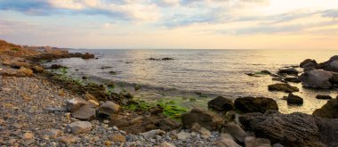 Taş ve çakıl taşlı deniz plajı. Güneşin doğuşunda gökyüzünde bulutlar olan güzel bir manzara. Bir körfezin geniş panoramik görüntüsü