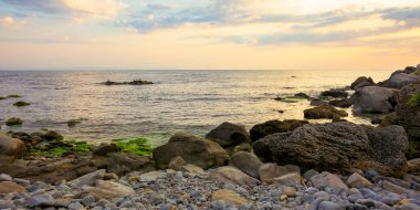 Taş ve çakıl taşlı deniz plajı. Güneşin doğuşunda gökyüzünde bulutlar olan güzel bir manzara. Bir körfezin geniş panoramik görüntüsü
