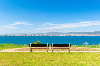 Güneşli sahil kıyısında iki bank. Sahildeki kaldırımlı patikadan güzel bir manzara. Şehir ve dağ, bulutlu mavi gökyüzünün altında.