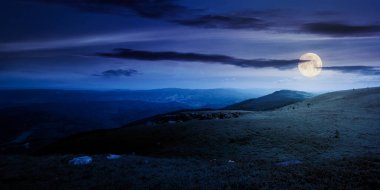 Çimenli alp çayırındaki kayalıklı çayır geceleri. Yazın dolunay ışığında dağ manzarası. Uzaktaki tepenin üstündeki mavi gökyüzünde bulutlar