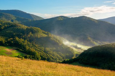 Akşamları dağlık kırsal alan. Ormandaki tepelerdeki tarlalar. Vadide gün ışığında parlayan duman ya da sis. Mavi gökyüzündeki bulutlar