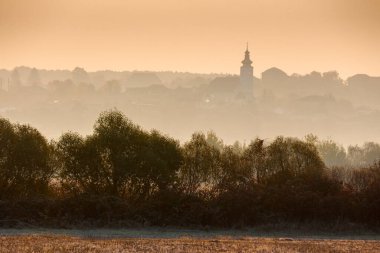 Kırsal alan bir sonbahar sabahında. Boş tarlalar boyunca sıra sıra ağaçlar. Tepede kilisesi olan bir köy ormanın arkasında sisli bir mesafede.