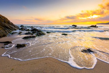 Sahilde gün doğumu. Deniz tatili için güzel bir yaz manzarası. Kumda kayalar. Sudaki sakin dalgalar. Gökyüzündeki bulutların arkasında güneş. Sozopol 'daki bambu plajının geniş panoramik manzarası