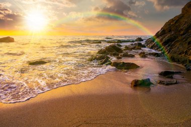 Gün batımında körfezdeki kayaların üzerinde gökkuşağı olan deniz kıyısı. Deniz gelgiti ve sakin dalgalar akşam ışığında altın kumlu sahili yıkıyor. Balayı için deniz kenarında güzel bir sonbahar tatili.