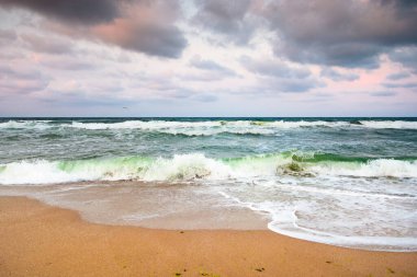 Gökyüzündeki manzaralı bulutların altında dalgaların kumsala vurduğu fırtınalı sahil manzarası. Yolculuk için karanlık, kasvetli bir arka plan. Avrupa kıyılarının manzarası
