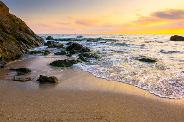 Sahilde gün doğumu. Deniz tatili için güzel bir yaz manzarası. Kumda kayalar. Sudaki sakin dalgalar. Gökyüzündeki bulutların arkasında güneş. Sozopol 'daki bambu plajının geniş panoramik manzarası