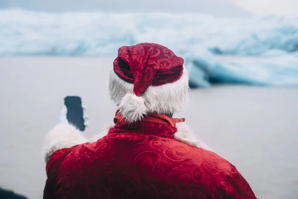 Traditional Style authentic Santa Claus on his back holding a mobile phone in his left hand in a glacier in front of a Iceland