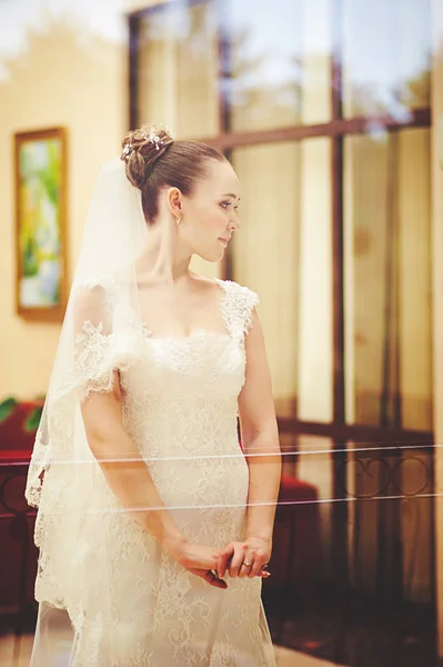 Bride in a white dress and a long veil in the hall behind the glass with reflection