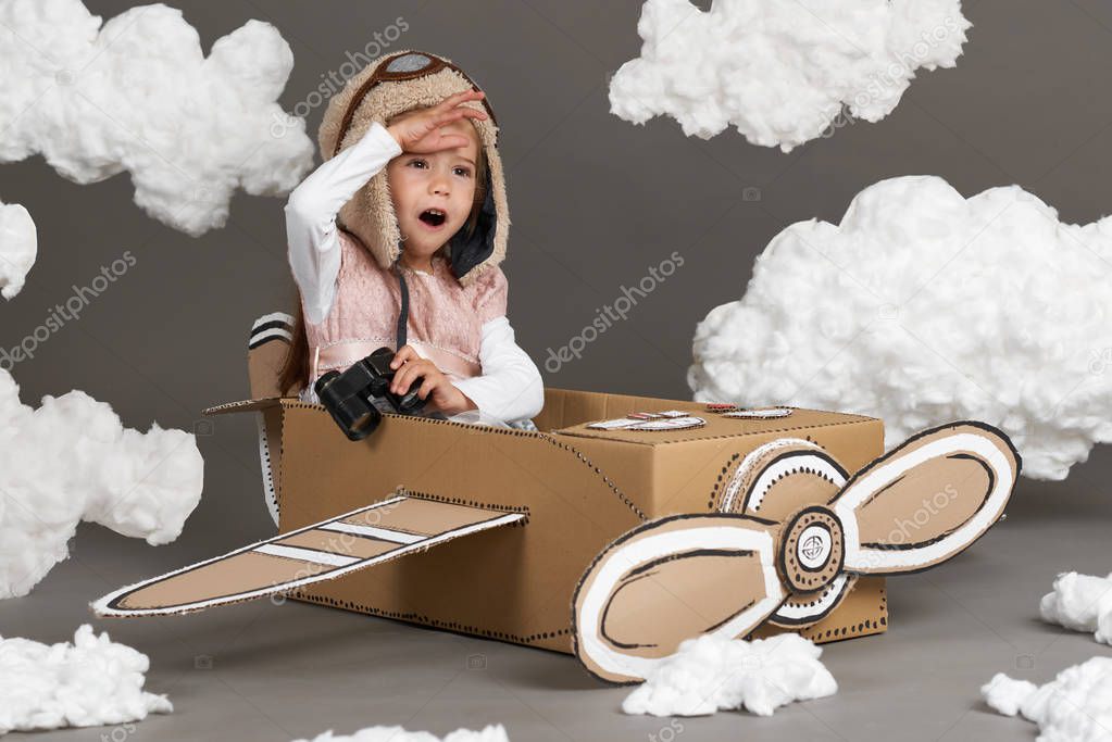 the child girl plays in an airplane made of cardboard box and dreams of becoming a pilot, clouds of cotton wool on a gray background