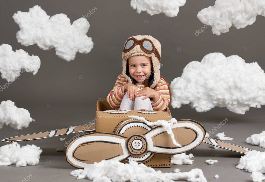 the child girl plays in an airplane made of cardboard box and dreams of becoming a pilot, clouds of cotton wool on a gray background