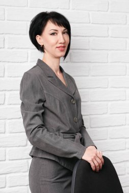 iş kadın giyinmiş bir gri takım elbise ile beyaz bir duvar önündeki bir sandalyeye pozlar