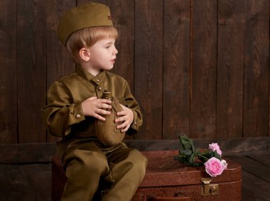 Çocuk Çocuk giymiş retro askeri üniformalı asker olarak oturan eski bavul, koyu ahşap arka plan, retro tarzı şişesi ile