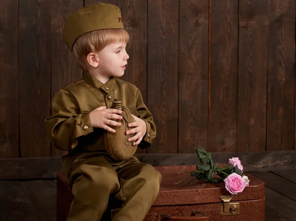 Çocuk Çocuk giymiş retro askeri üniformalı asker olarak oturan eski bavul, koyu ahşap arka plan, retro tarzı şişesi ile