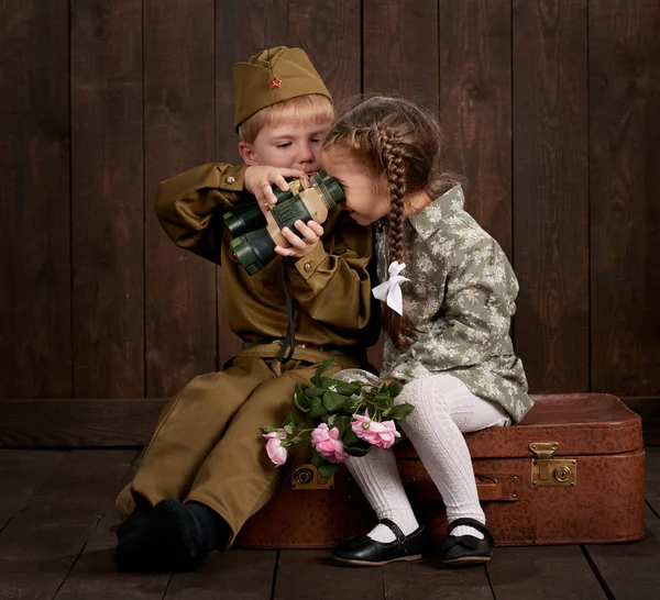Çocuk Çocuk retro askeri üniformalı asker ve kız pembe elbiseli oturma eski bavul, koyu ahşap arka plan, retro tarzı olarak giymiş