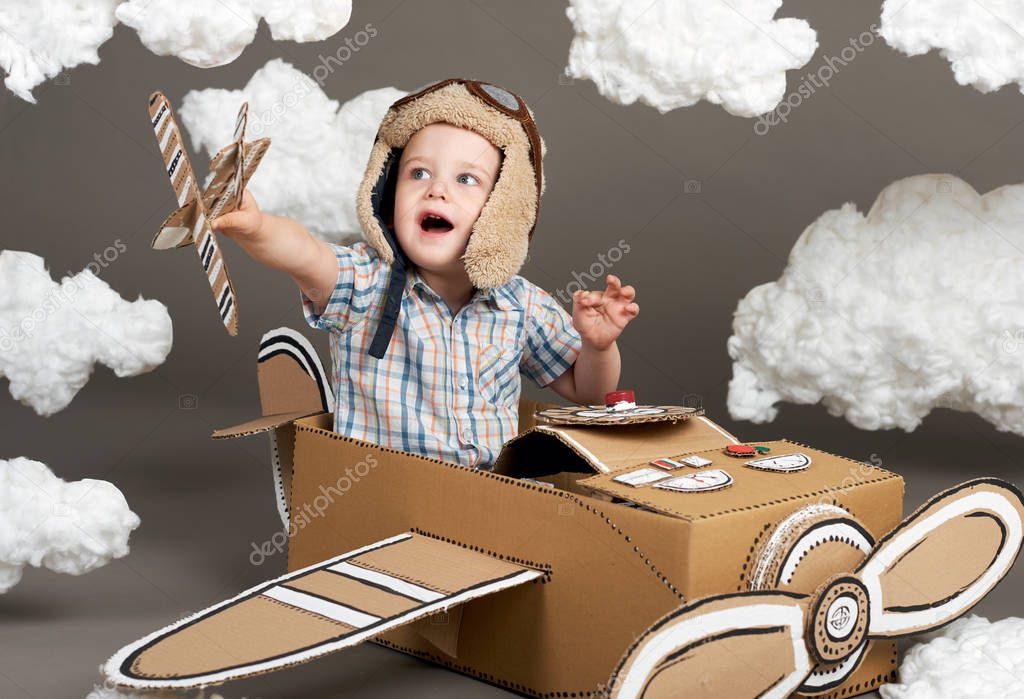 the boy plays in an airplane made of cardboard box and dreams of becoming a pilot, clouds from cotton wool on a gray background, retro style
