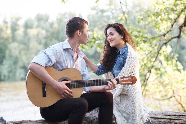 young couple sitting on a log by the river and playing guitar, summer nature, bright sunlight, shadows and green leaves, romantic feelings