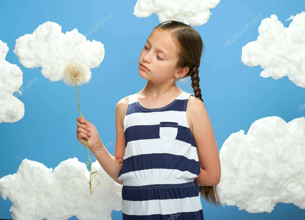 girl has a big dandelion in her hands, dressed in striped dress, posing on a blue background with cotton clouds, the concept of summer, holiday and happiness