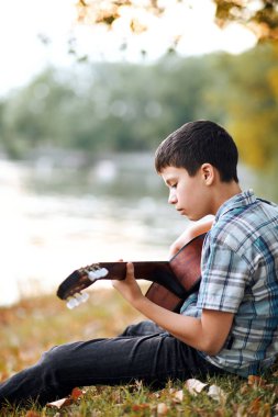 Çocuk akustik gitar çalar, nehir, gün batımı, güzel Doğa Sonbahar Ormanı ve ağaçlar su yansıması kıyısında oturuyor